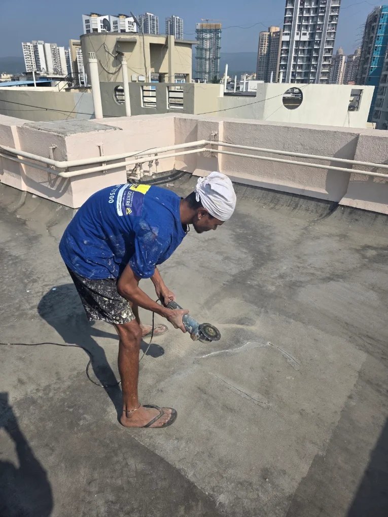 Worker angle-grinding a terrace surface to prep it for waterproofing, Mumbai skyline behind